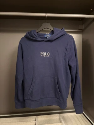 Marinblå hoodie från Polo Ralph Lauren - Snygg marinblå hoodie från Polo Ralph Lauren med klassisk logga broderad på bröstet. Hoodien har huva, känguruficka och ribbade muddar vid ärmar och nederkant. Perfekt för en avslappnad och stilren look.