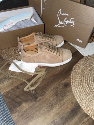 Beige sneakers från Christian Louboutin - Stilrena beige sneakers i mocka från Christian Louboutin med klassisk röd sula och vita kanter. Skorna har lågt skaft, tonade skosnören och diskreta sömmar. Perfekta för dig som vill ha en lyxig och trendig look.