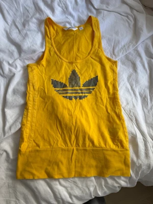 Gul linnetopp med Adidas-logga - Snygg gul linnetopp med stor Adidas-logga i svart på bröstet. Toppen är ärmlös och har en rund halsringning. Perfekt för dig som gillar sportig och färgglad stil.