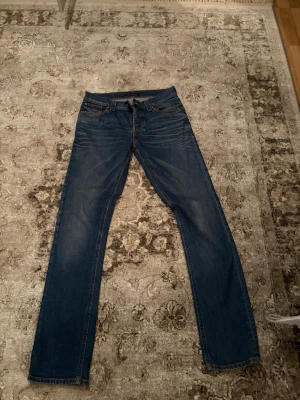 Mörkblå nudie jeans  - Säljer ett par mörkblå jeans med raka ben och snygga kontrastsömmar. Jeansen har klassisk femficksdesign och subtila slitningar framtill och baktill för en avslappnad look. Perfekta till vardagsstilen och passar till det mesta.