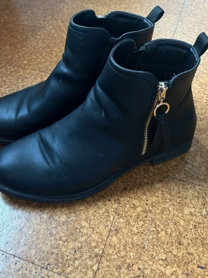 Svarta stövletter med dragkedja - Snygga svarta stövletter i matt skinnimitation med rund tå och dekorativ guldfärgad dragkedja på sidan. Klassisk modell med låg klack och ögla bak för enkel påtagning. Perfekta till jeans eller kjol för en stilren look.