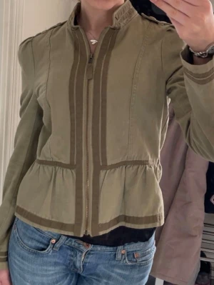 Grön jacka med dragkedja och detaljer - Snygg grön jacka med markerade sömmar och dekorativa band i mörkare grön. Jackan har hög krage och dragkedja framtill. Perfekt för dig som vill ha en unik och trendig look.