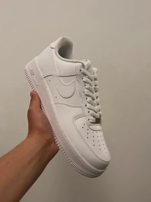 Vita Nike Air Force 1 sneakers - Klassiska helvita Nike Air Force 1 sneakers med låg profil. Skorna har ovandel i läder, perforerad tå för ventilation och tydlig Nike Swoosh på sidorna. Tjock sula med AIR-dämpning för extra komfort. Perfekta för en clean och stilren look.