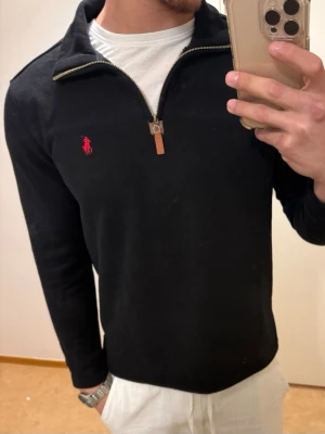 Svart stickad tröja från Ralph Lauren - Snygg svart stickad tröja från Ralph Lauren med halv dragkedja och klassisk röd logga på bröstet. Tröjan har lång ärm, ribbade muddar och en stilren look som passar till det mesta. Perfekt för dig som gillar tidlös och enkel stil.