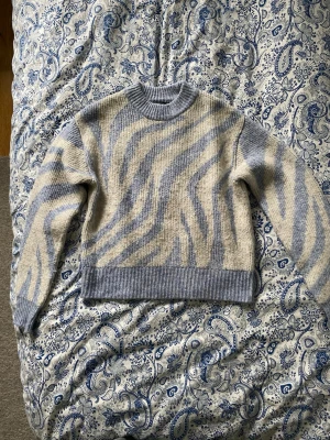 Stickad tröja med zebramönster  - Säljer en stickad tröja från Gina Tricot. Tröjan har ett zebramönster i ljusblått och krämvitt, med ribbade muddar vid hals, ärmslut och nederkant. Jag har inte använt tröjan mer än 2-3 gånger. 