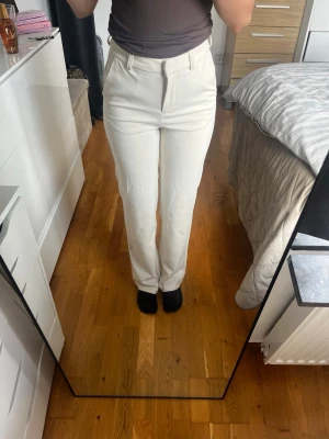 Vita raka kostymbyxor - Säljer ett par stilrena vita kostymbyxor med rak passform och hög midja. Byxorna har bälteshällor och diskreta fickor framtill. Perfekta för en clean och modern look. Materialet ser ut att vara en mjuk och lätt blandning, vilket ger en snygg siluett.