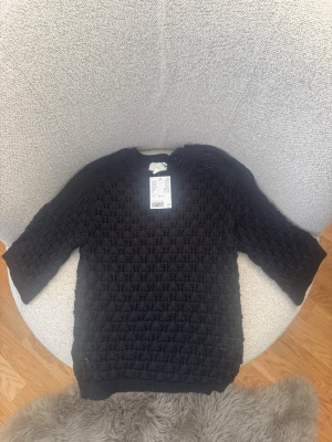 Svart stickad tröja från H&M - Snygg svart stickad tröja från H&M med strukturmönster och rund hals. Tröjan har ribbade muddar vid ärmslut och nederkant samt en klassisk passform. Perfekt att matcha med jeans eller kjol för en trendig look.