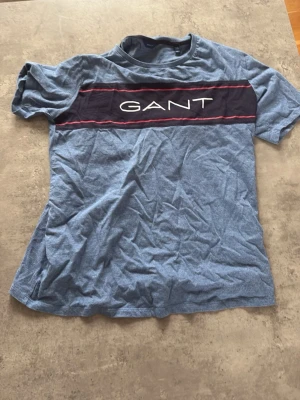 Blå GANT t-shirt med logga - Säljer en blå t-shirt från GANT med korta ärmar och en bred mörkblå rand över bröstet där GANT-loggan är tryckt i vitt. T-shirten har en klassisk passform och är gjord i mjuk bomull. Perfekt till vardags för en avslappnad stil. Storlek 176cm=S 
