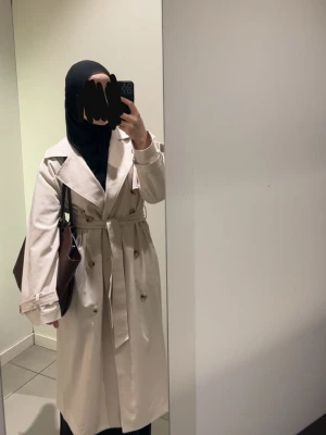 Beige klassisk trenchcoat med skärp - Stilren beige trenchcoat med dubbelknäppning, breda slag och knytskärp i midjan. Jackan har långa ärmar med justerbara remmar vid ärmsluten och två sidofickor. Perfekt för lager-på-lager och ger en tidlös look. S men är oversize 