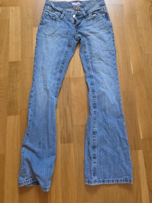 Ljusblå bootcut jeans låg midja - Säljer ett par ljusblå bootcut jeans med låg midja och klassiska fem fickor. Jeansen har en avslappnad passform och är tillverkade i denim. Perfekta för dig som gillar en retro vibe och vill ha ett par jeans som sticker ut.