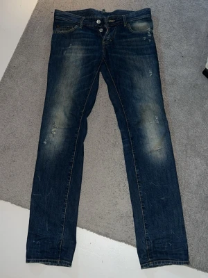 Dsquared2 jeans slitningar - Säljer ett par mörkblå jeans med slitningar och snyggt tvättade partier på låren och knäna. Jeansen har klassisk femficksdesign, knappgylf och smal passform. Perfekta för en avslappnad och trendig look. Ärvda från en i familjen köpta i USA. Jätte fint skick på dom. Storlek 46, i eu är de storlek ca 29-30 passar de. L32