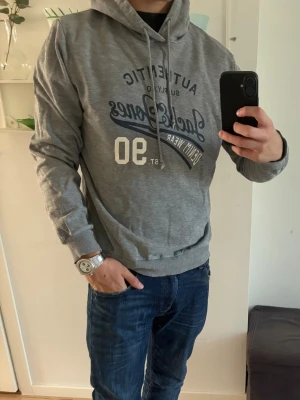  Jack & Jones - Snygg grå hoodie från Jack & Jones med tryck i blått och vitt på bröstet. Klassisk huva med dragsko och ribbade muddar vid ärmar och midja. Perfekt för en avslappnad stil och enkel att matcha med jeans.