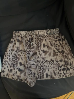 Kort leopardmönstrad kjol med volang - Säljer en kort kjol med leopardmönster i beige och svart. Kjolen har en elastisk midja och en snygg volangdetalj framtill som ger extra rörelse. Perfekt för dig som vill sticka ut med ett djuriskt print! Den är i storlek L men jag skulle själv säga att den passar bäst till S/M. Kjolen har även inbyggda shorts i sig!💞