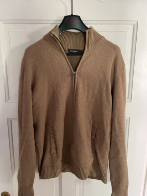 Beige ziptröja från Dressmann - Beige stickad ziptröja från Dressmann med hög krage och ribbade muddar. Tröjan har två fickor framtill och dragkedja hela vägen. Perfekt för lager-på-lager och enkel att matcha med olika outfits.