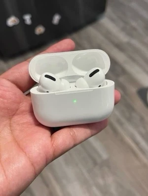 Apple AirPods Pro  - A hand is holding a pair of Apple AirPods Pro inside their white charging case. The case is open, and a green LED light is visible on the front, indicating that the earbuds are charged or charging. The background shows a wooden floor and part of a dark object with stickers.
