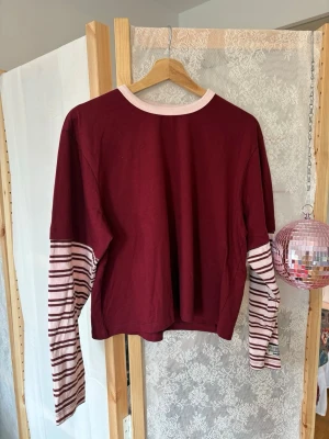 Vinröd långärmad topp med ränder - Vinröd långärmad topp med kontrasterande rosa och vinröda randiga ärmar samt ljusrosa halsringning. Toppen har en loose passform och är tillverkad i mjuk bomull, perfekt för en avslappnad stil.