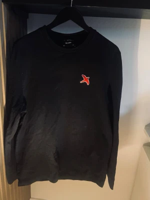 Svart långärmad tröja från Axel Arigato - En stilren svart långärmad tröja från Axel Arigato med den Klassiska röda fågelbrodyr på bröstet. Tröjan har enkel design och är gjord i mjukt material, perfekt för en clean och avslappnad look. Storlek S men passar M, pris kan diskuteras vid snabb affär