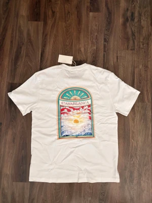 Vit Casablanca t-shirt med tryck - Vit t-shirt från Casablanca med färgglatt tryck på ryggen som föreställer ett fönster med soluppgång och texten 'CASABLANCA'. På bröstet finns ett mindre tryck med en snäcka och logotyp. Klassisk passform och rund hals.