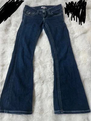 Mörkblå bootcut jeans med kontrastsömmar - Säljer ett par mörkblå bootcut jeans med vita kontrastsömmar och låg midja. Jeansen har klassiska fem fickor och utsvängda ben, perfekta för en retroinspirerad look. Materialet är denim i bomull som känns slitstarkt och bekvämt.