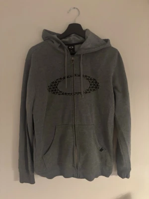 Grå oakley hoodie - Grå hoodie med dragkedja framtill och svart tryck på bröstet. Tröjan har huva med snörning och två fickor fram. Tillverkad i mjukt bomullsmaterial, perfekt för chill dagar. Klassisk passform och enkel design.