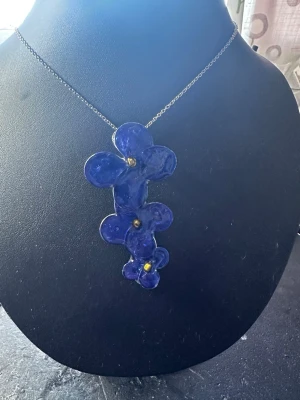 Blått halsband med blommor - Unikt halsband med hängsmycke i form av tre blå blommor i olika storlekar, dekorerade med små guldfärgade detaljer i mitten. Kedjan är silverfärgad och smycket har en konstnärlig, handgjord känsla. Perfekt för dig som gillar statement-smycken och färg.
