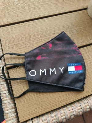 Svart ansiktsmask Tommy Hilfiger - Snygg svart ansiktsmask med Tommy Hilfiger-logga och diskreta röda detaljer. Masken har elastiska öronband och är tillverkad i ett mjukt tygmaterial. Perfekt accessoar för dig som vill ha både stil och funktion. Knappt använd och tvättad 
