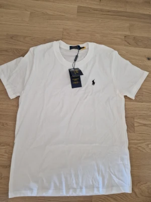Ralph Lauren t-shirt - Klassisk vit t-shirt från Polo Ralph Lauren med rund halsringning och ikoniska logobroderiet på bröstet. Tillverkad i mjuk bomull och har en normal passform. Perfekt basplagg för en clean och stilren look.
