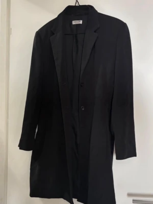 Svart klassisk kappa med knappar - Stilren svart kappa med rak passform och klassisk krage. Kappan har två knappar framtill, lätta axelvaddar och långa ärmar. Perfekt för dig som gillar en minimalistisk och tidlös look. Passar bra till både jeans och kostymbyxor.