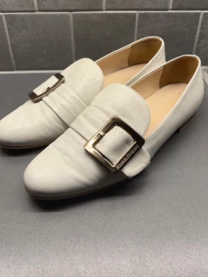 Cremefärgade loafers från Russell & Bromley - Stilrena vita loafers från Russell & Bromley i mjukt läder. Skorna har en fyrkantig tå och ett stort, silverfärgat metallspänne med märkets logga på ovansidan. Låg klack och klassisk design som passar till många olika looks. Lätt slitage på lädret på framsidan (se bild) samt vagt märke från spännet. 