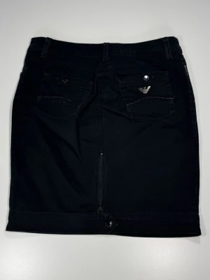 Armani Jeans Svart kortkjol med fickor - Snygg svart kortkjol med två bakfickor, varav ena har en liten metallogo. Kjolen har bälteshällor, knapp och dragkedja framtill samt en liten slits bak. Perfekt till en avslappnad eller trendig look.0091