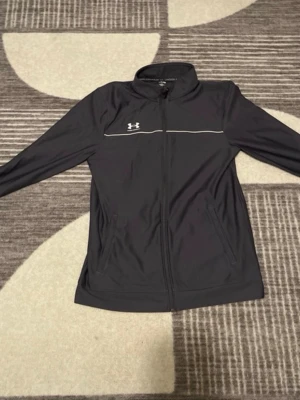 En äkta underarmour tröja helt ny köpte den från Jd sports  - Snygg svart träningsjacka från Under Armour med hel dragkedja framtill och diskret logga på bröstet. Jackan har ståkrage, två sidofickor och en tunn vit linje över bröstet för extra stil. Perfekt för gymmet eller löprundan.