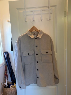Beige overshirt från Jack & Jones - Stilren beige overshirt från Jack & Jones med två stora bröstfickor och svarta knappar. Overshirten har klassisk krage och är tillverkad i ett mjukt, tjockare tyg som passar perfekt för lager-på-lager. Enkel att matcha med olika outfits.