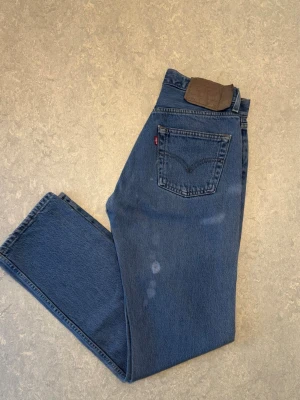 Blå raka Levis 501 - Säljer ett par klassiska blå jeans med rak passform och hög midja. Jeansen har en bakficka med dekorativ söm och en liten röd etikett. Tillverkade i kraftigt denimtyg som ger en tidlös look. Allt förutom Levis etiketten är i väldigt bra skick. Säljer de pågrund av att de är för små. Passar allt och har en tidlös design som alltid kommer vara trendig