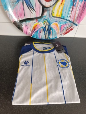 Bosnien & Hercegovina matchtröja (Kelme) – Helt ny med lappar! - Tröjan har en riktigt snygg design i vitt, blått och gult med finstämda ränder och förbundsmärket på bröstet. 🔥 Passa på att köpa nu i god tid innan landskampen mellan Sverige och Bosnien på Strawberry Arena! Perfekt att ha på sig på läktaren för att visa ditt stöd. Har fler storlekar,hör av dig pm.