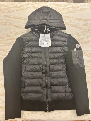Svart pufferjacka med huva - Säljer en svart pufferjacka med stickade ärmar och huva. Jackan har quiltad front ifrån moncler, dragkedja framtill och en ficka med dragkedja på ena ärmen. Moncler-logga på ärmen och ribbade muddar. Perfekt för kalla vinterdagar och riktigt snygg streetstil.