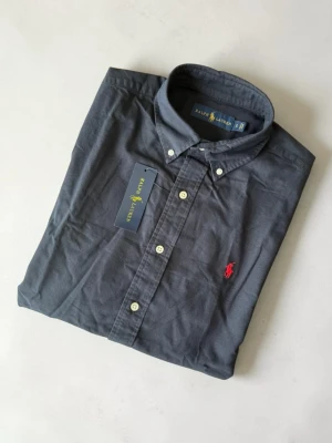 Mörkblå skjorta från Ralph Lauren - Klassisk mörkblå skjorta från Ralph Lauren med button-down krage och röd broderad logga på bröstet. Skjortan har långa ärmar, vita knappar och är tillverkad i mjuk bomull. Perfekt för en stilren och avslappnad look.