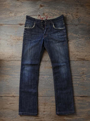 Pepe jeans - Mörkblå jeans från Pepe Jeans i fint skick Storlek W33 L34  Midjemått: ca 42 cm Innerbenslängd: ca 83 cm Mörkblå denim med snygga slitningar/detaljer Bra skick, inga större synliga defekter Knäppning med knappar framtill