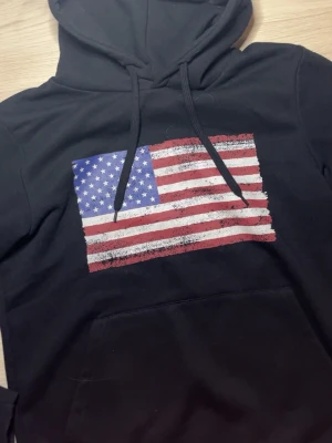 Svart hoodie med amerikansk flagga - Säljer en svart hoodie med stor amerikansk flagga tryckt på bröstet. Hoodien har huva med dragsko och en klassisk magficka. Perfekt för dig som gillar streetwear och vill sticka ut med en cool print.