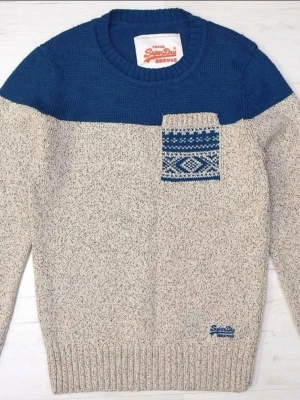 Beige och blå stickad tröja Superdry - Stickad tröja från Superdry i beige och blått med rund halsringning. Tröjan har en blå överdel och en beige kropp, samt en liten bröstficka med blått och vitt mönster. Ribbstickade muddar vid ärmslut och nederkant.