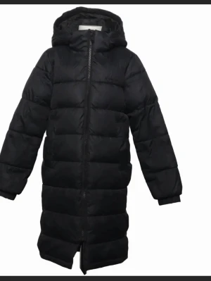 Svart lång jacka vinter jacka  - Snygg svart lång dunjacka med huva och dragkedja framtill. Jackan har quiltad design med breda kanaler och ribbade muddar vid ärmsluten. Perfekt för kalla vinterdagar och ger en stilren look.