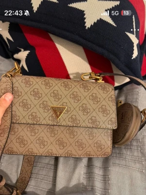 Beige monogram handväska från Guess - Snygg beige handväska från Guess med klassiskt monogrammönster och guldfärgade detaljer. Väskan har en justerbar axelrem och en liten rund coin pouch som hänger på sidan. Perfekt för dig som gillar stilrena accessoarer.