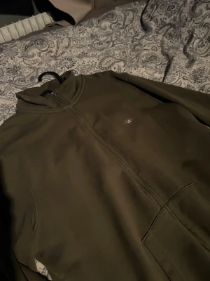 Gant zip hoodie - Olivgrön ziptröja med hög krage och två framfickor. Liten broderad detalj på bröstet. Perfekt för lager-på-lager och chill dagar. Mjuk och bekväm bomullskänsla.