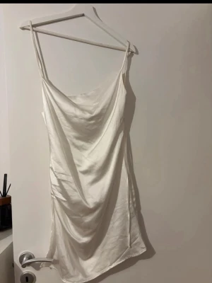 Vit sidenklänning med tunna band - Säljer en elegant vit klänning i sidenlook med tunna axelband och draperad front. Klänningen är kort och har en mjuk, glansig yta som ger en lyxig känsla. Perfekt för dig som gillar minimalistisk och stilren design.