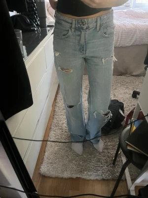 Ljusblå slitna raka jeans - Säljer ett par ljusblå jeans med raka ben och flera slitningar och hål på både fram- och baksidan. Jeansen har klassisk femficksmodell och hög midja. Perfekta för en avslappnad och trendig look.