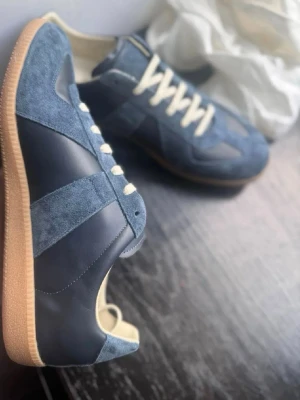 Blå Maison Margiela sneakers - Snygga sneakers från Maison Margiela i blå mocka och läder med vita snören och klassisk gummisula. Skorna har en låg silhuett och retroinspirerad design som passar perfekt till streetwear och casual outfits. Skorna är använda 1 gång, verkligen toppskick. Storlek 44. Kommer utan box då jag köpt dessa på andra hand.