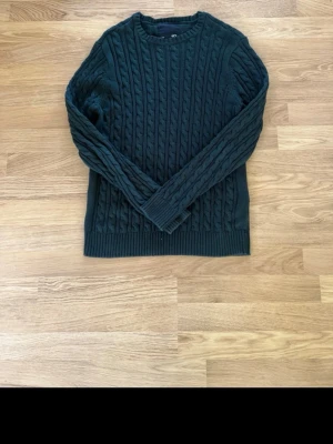 Mörkgrön kabelstickad tröja - Snygg mörkgrön kabelstickad tröja med rund halsringning och långa ärmar. Tröjan har ett klassiskt flätmönster över hela plagget och ribbade muddar vid ärmslut och nederkant. Perfekt för dig som gillar en stilren och mysig look.