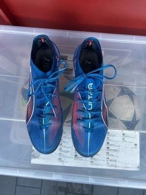 Blåa Puma ultra elite fotbollsskor med dobbar - Säljer ett par blåa fotbollsskor från Puma med vita och röda detaljer. Skorna har snörning och är tillverkade i syntetmaterial. Perfekta för spel på konstgräs med sina fasta dobbar och sportig design. Skorna är använda 5 gånger. Puma ultra påse ingår ej då den va sönder när jag fick skorna!
