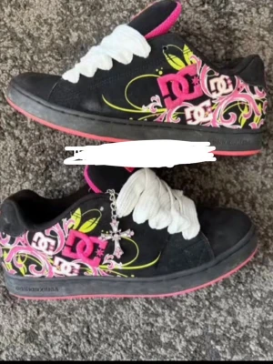 SÖKER!!!! - Säljer ett par svarta DC sneakers med stora rosa och vita loggor på sidan, samt färgglada grafiska mönster i gult och rosa. Skorna har breda vita skosnören, rosa insida och rosa sula. Perfekta för dig som gillar streetwear och unika detaljer.