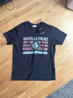 Svart Saint Laurent t-shirt med tryck - Säljer en svart t-shirt från Saint Laurent med coolt tryck framtill i vitt och rött. T-shirten har korta ärmar och klassisk passform. Perfekt för dig som vill ha en stilren men ändå statement look.