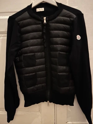 Svart quiltad cardiganjacka Moncler - Svart cardiganjacka från Moncler med quiltad framsida och två fickor. Jackan har dragkedja framtill, rund halsringning och Moncler-logga på ärmen. Ärmar och rygg är i stickat material medan framsidan är vadderad för extra värme. Skrev för mer information priset går att diskutera vid snabb affär 